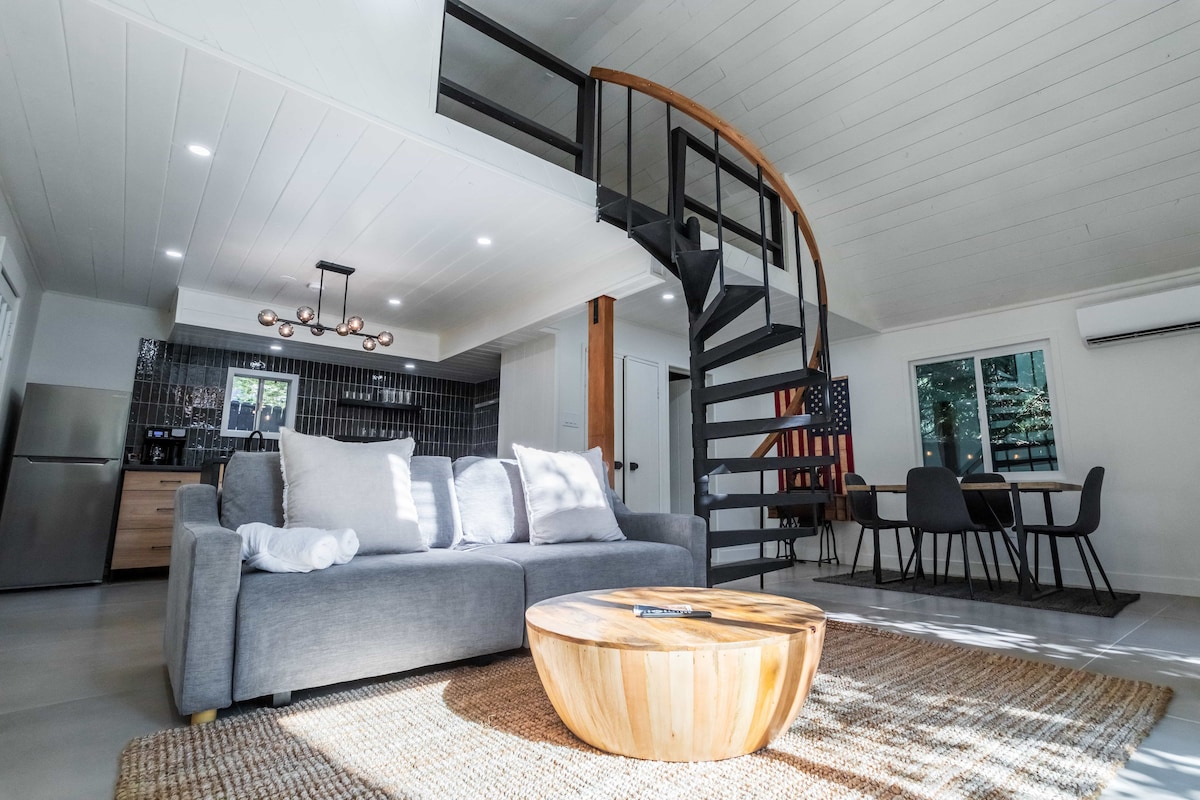 A spacious living area is showcased, featuring a comfortable sofa and a round wooden coffee table. A spiral staircase leads to the loft, while the kitchen area is visible in the background, along with a dining table that accommodates four. Natural light floods the space through large windows.