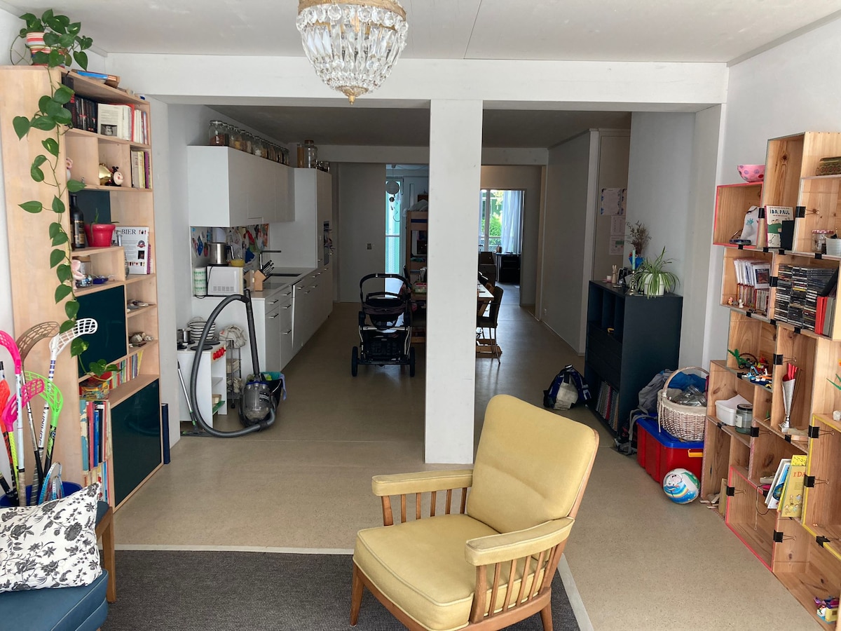 An open living space is featured, with a bright yellow armchair positioned on a dark rug. A modern kitchen with white cabinetry is visible, alongside a dining area with wooden chairs. Natural light filters in through central glass doors, while various shelves are stocked with books and decor.