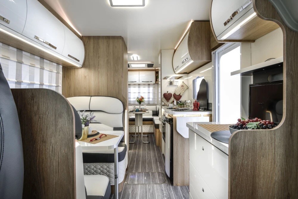 The interior features a modern layout with sleek cabinetry and earth-toned finishes. A dining area includes a table surrounded by cushioned seating. A well-equipped kitchen is visible, showcasing appliances and countertops, complemented by natural light entering through a nearby window.