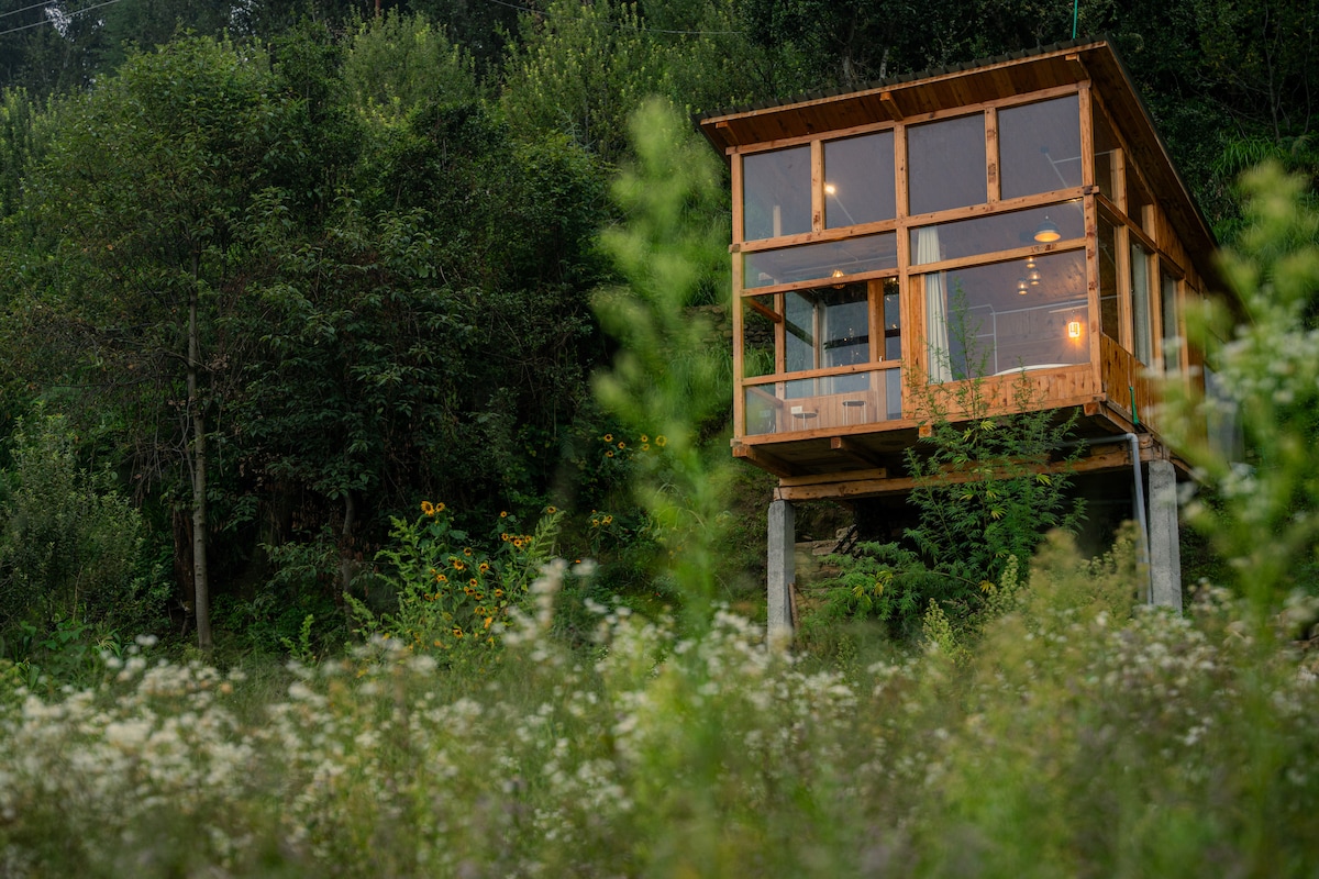 A modern treehouse is elevated on stilts, surrounded by lush greenery and wildflower fields. Its wooden structure features expansive glass walls, allowing natural light to flood the interior while providing views of the tranquil landscape.