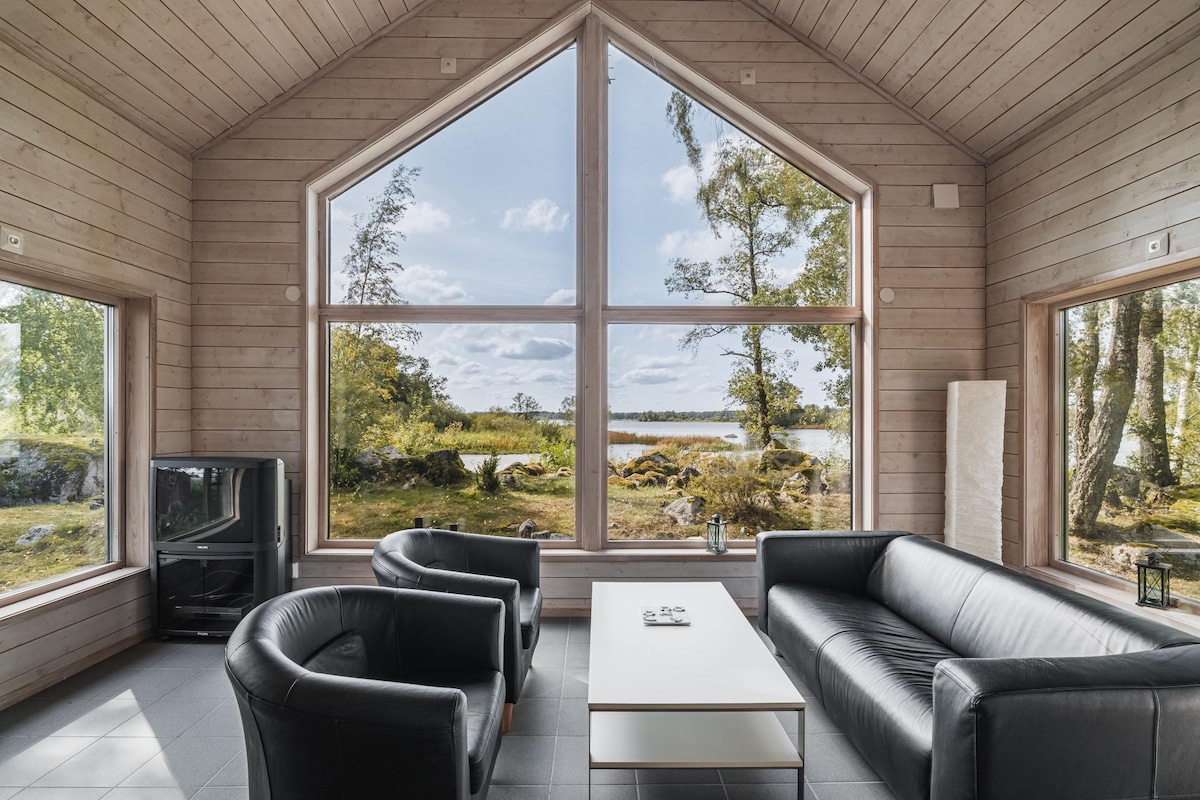 A bright living area is characterized by large triangular windows, providing a view of the surrounding nature. Two black armchairs and a black sofa are arranged around a white coffee table. A sleek stove is positioned in one corner, enhancing the minimalist decor.
