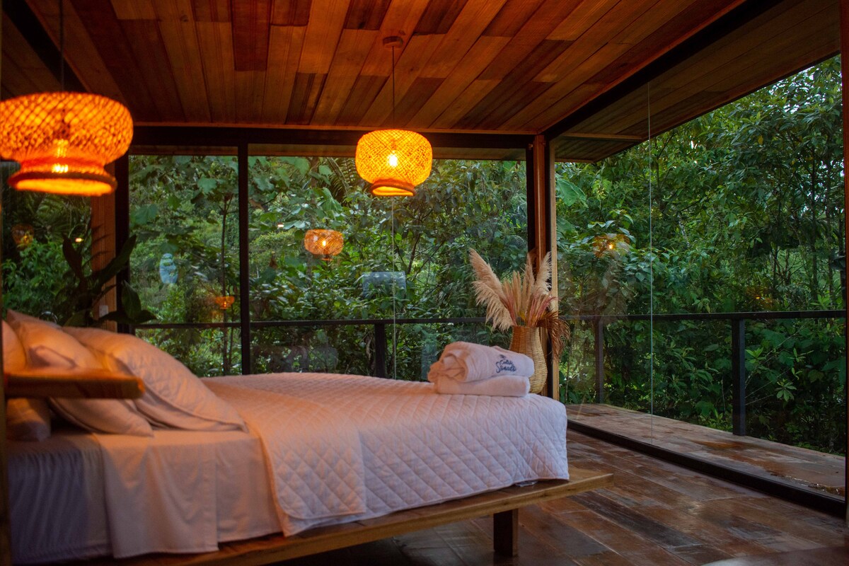 A spacious bed with white bedding is situated in a glass-walled room, allowing for views of the surrounding greenery. Warm lighting is provided by woven pendant lamps, creating a serene atmosphere. Natural elements, such as dried plants, enhance the connection to the outdoor environment.