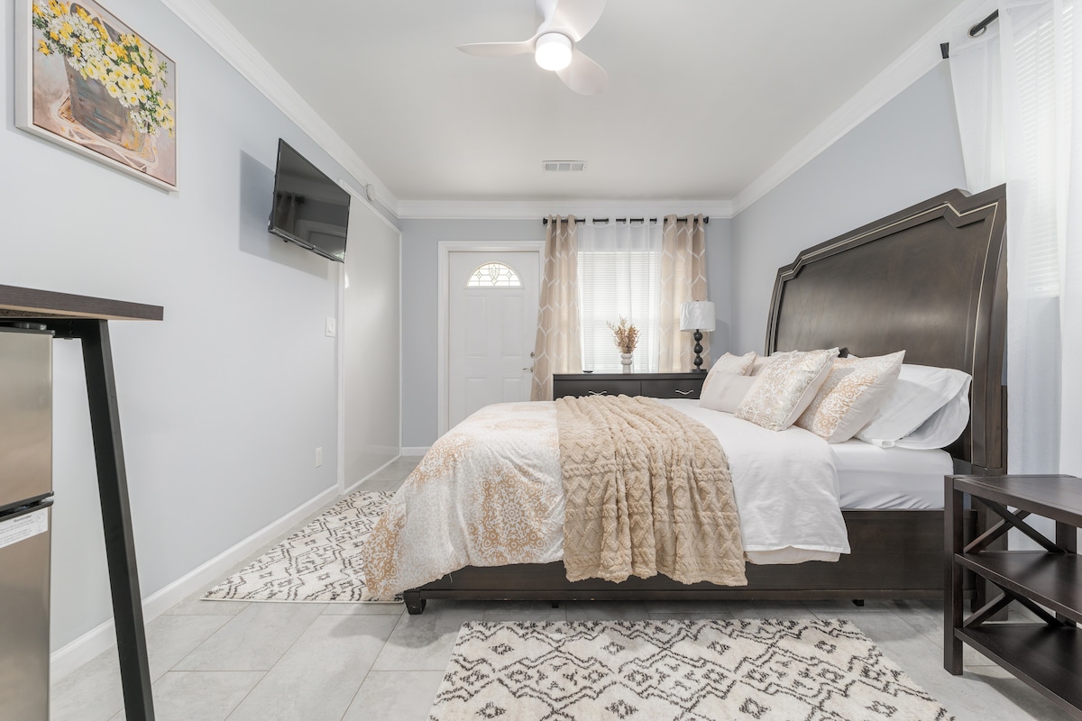 A private room features a queen bed with decorative pillows and a textured throw blanket. A small table and a side shelf are positioned beside the bed. Natural light enters through sheer curtains covering the window, and a flat-screen TV is mounted on the wall.