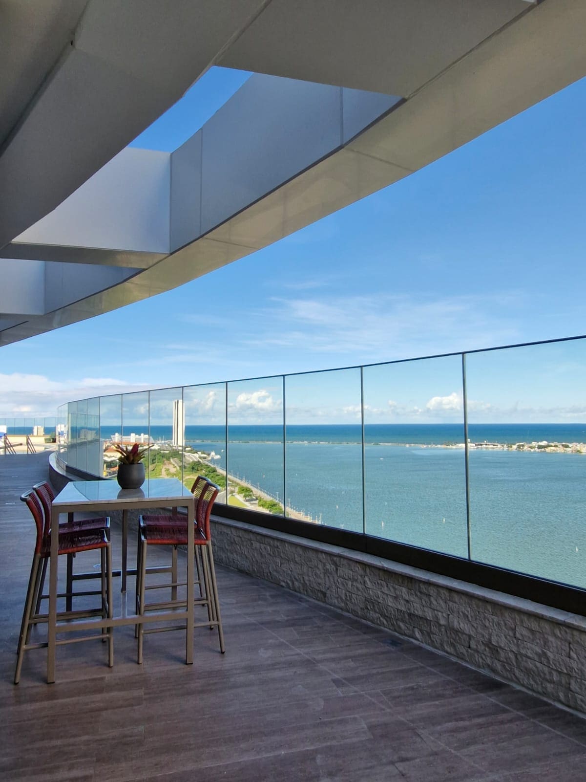 A spacious balcony features a table with high chairs, providing views of the water and the surrounding landscape. The glass railing allows for unobstructed sightlines, while the architectural design offers shelter and modern aesthetics.