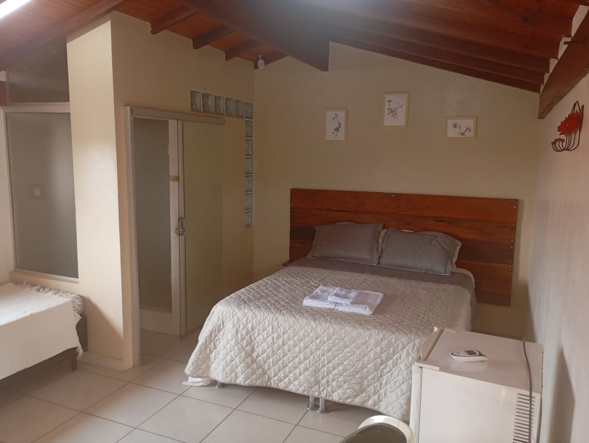 A cozy bedroom features a spacious bed adorned with smooth linens. A single bed is placed in the corner. The room is complemented by wooden beams overhead and light-colored walls, with a small round table and a fridge present for convenience.