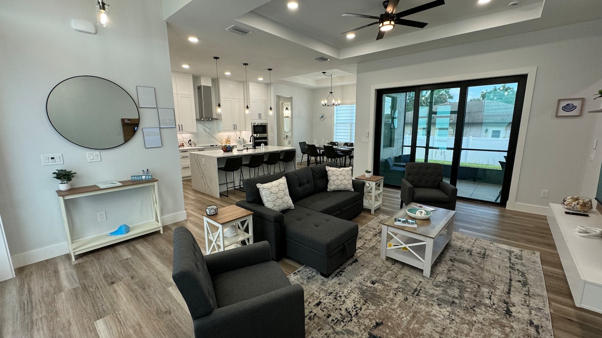 A modern living area features a sectional sofa and two accent chairs arranged around a coffee table. Light wood flooring complements the neutral color palette. A large mirror reflects the room's brightness, while sliding glass doors offer views of the screened lanai.