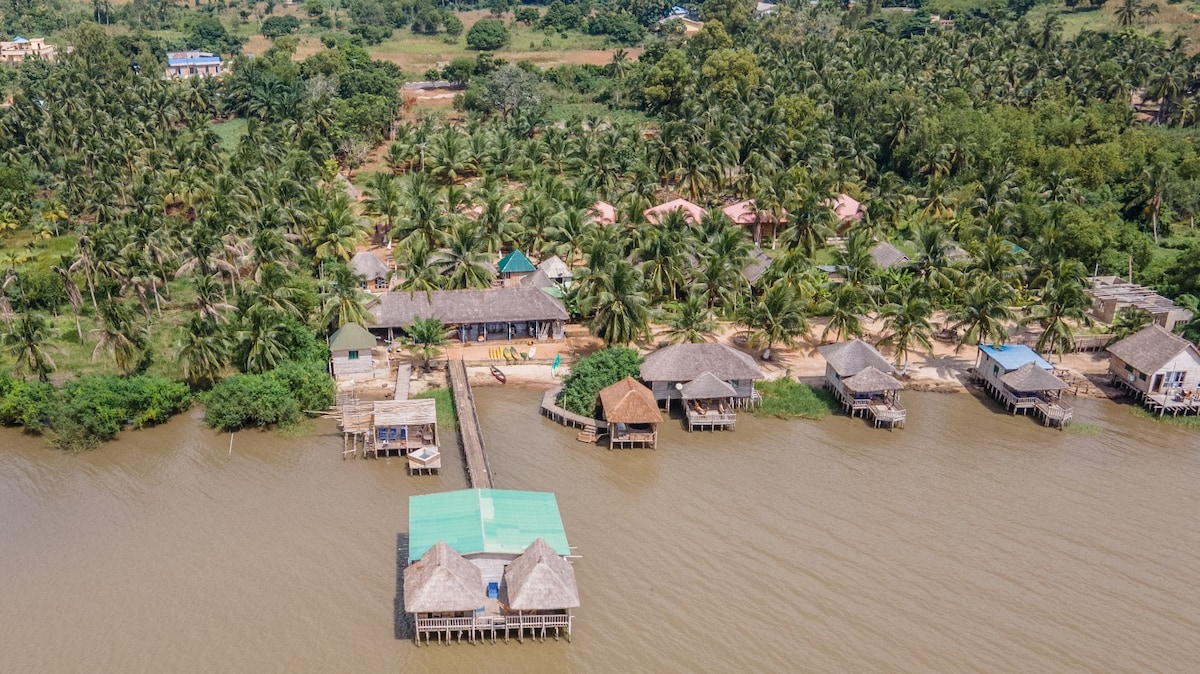 An aerial view captures a collection of thatched-roof and wooden structures along a riverbank, surrounded by lush greenery. A wooden walkway connects the buildings to the water, highlighting a serene and natural environment.