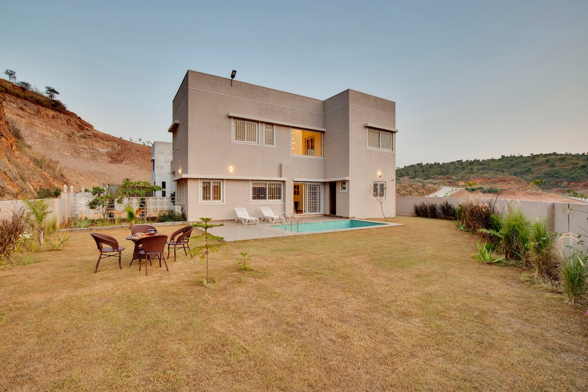 The villa features a modern exterior with a two-story structure. A private outdoor pool and loungers are seen in the foreground, surrounded by a spacious grassy area. Natural hills can be observed in the background, enhancing the serene environment.