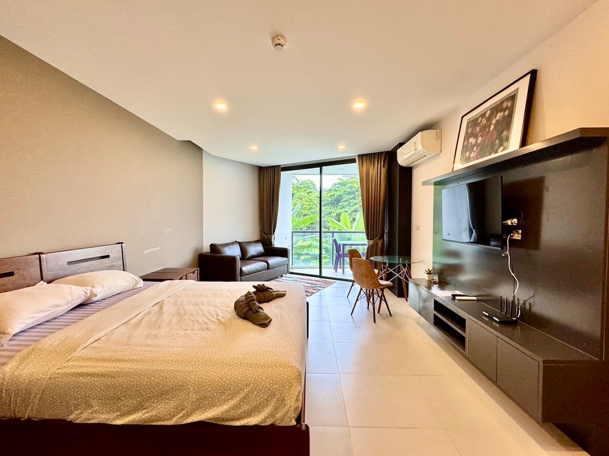 A spacious room features a large bed with light bedding, accompanied by a brown sofa and a glass dining table. Natural light enters through floor-to-ceiling windows, revealing a view of greenery outside. A mounted TV is positioned on the wall, enhancing the functionality of the space.
