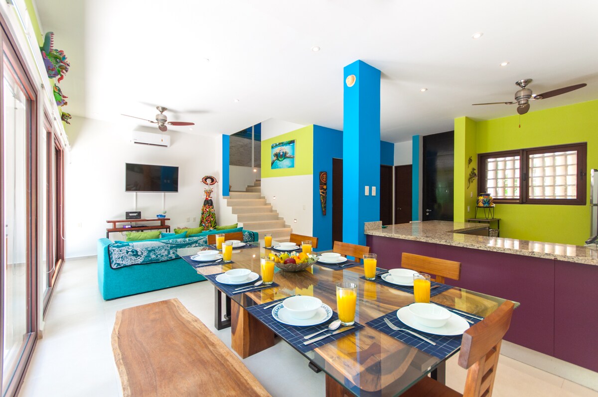 An open-concept living space is shown, featuring a dining table set for breakfast with orange drinks and plates. A vibrant blue and green color scheme highlights the modern design. A cozy seating area with a large couch is visible, alongside a kitchen area with granite countertops.
