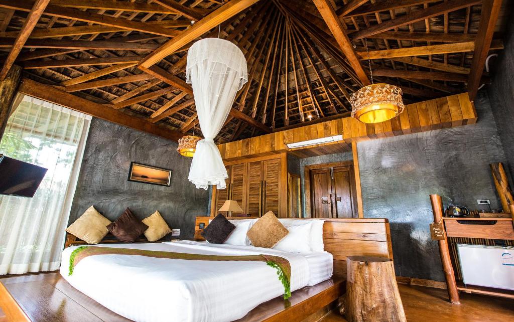 A spacious room features a large wooden bed adorned with decorative pillows. A draped canopy adds a touch of elegance to the high ceiling, made of dark wood. Natural light pours in through large windows, complemented by wooden furnishings and rustic accents.