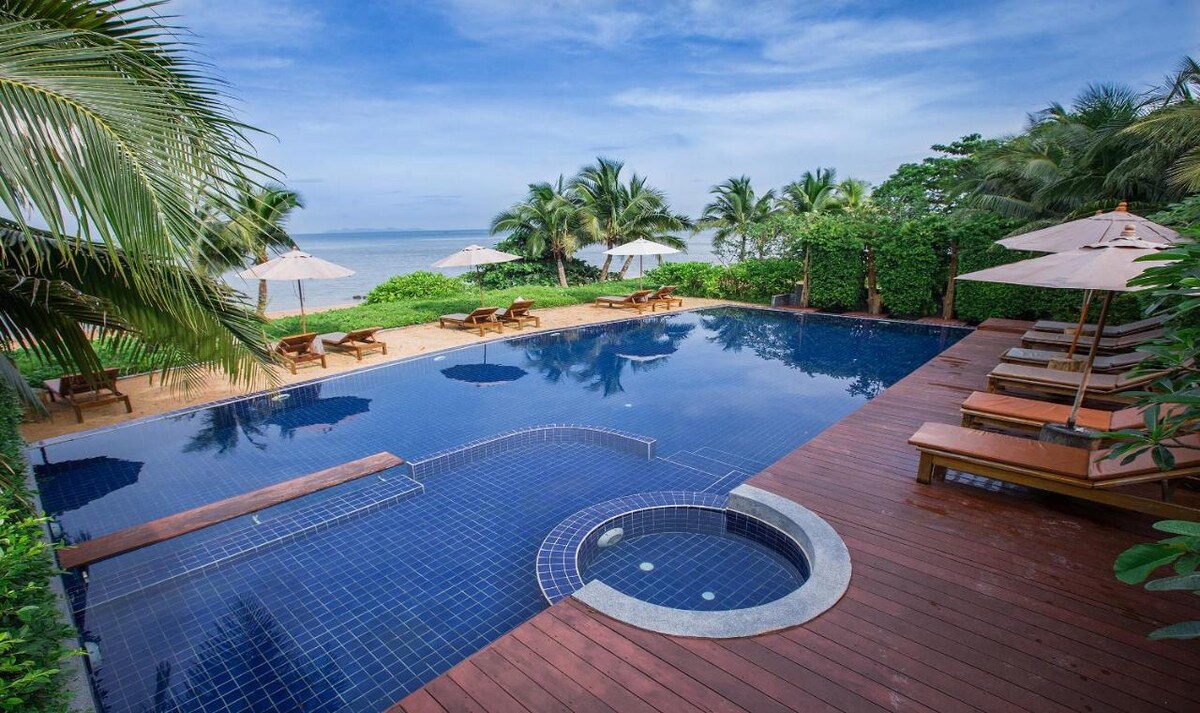An outdoor pool is surrounded by a wooden deck, featuring lounge chairs and umbrellas along its perimeter. Lush greenery frames the area, and an inviting hot tub is integrated into the pool's design. The serene ocean view complements the tropical setting.