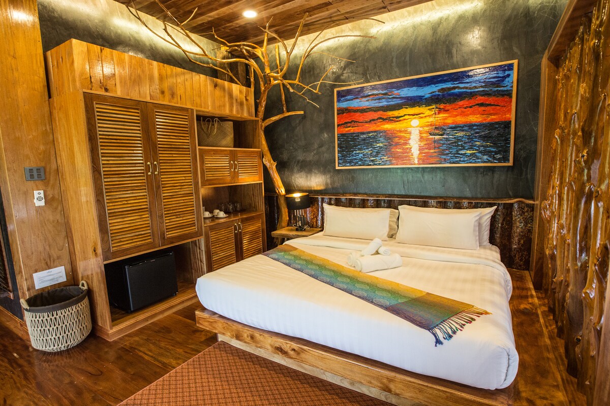 A spacious sleeping area is highlighted by a wooden bed frame and a soft white duvet. A colorful blanket lays diagonally across the bed. An artwork featuring a sunset adorns the wall above. A wooden cabinet and a small refrigerator are also present.