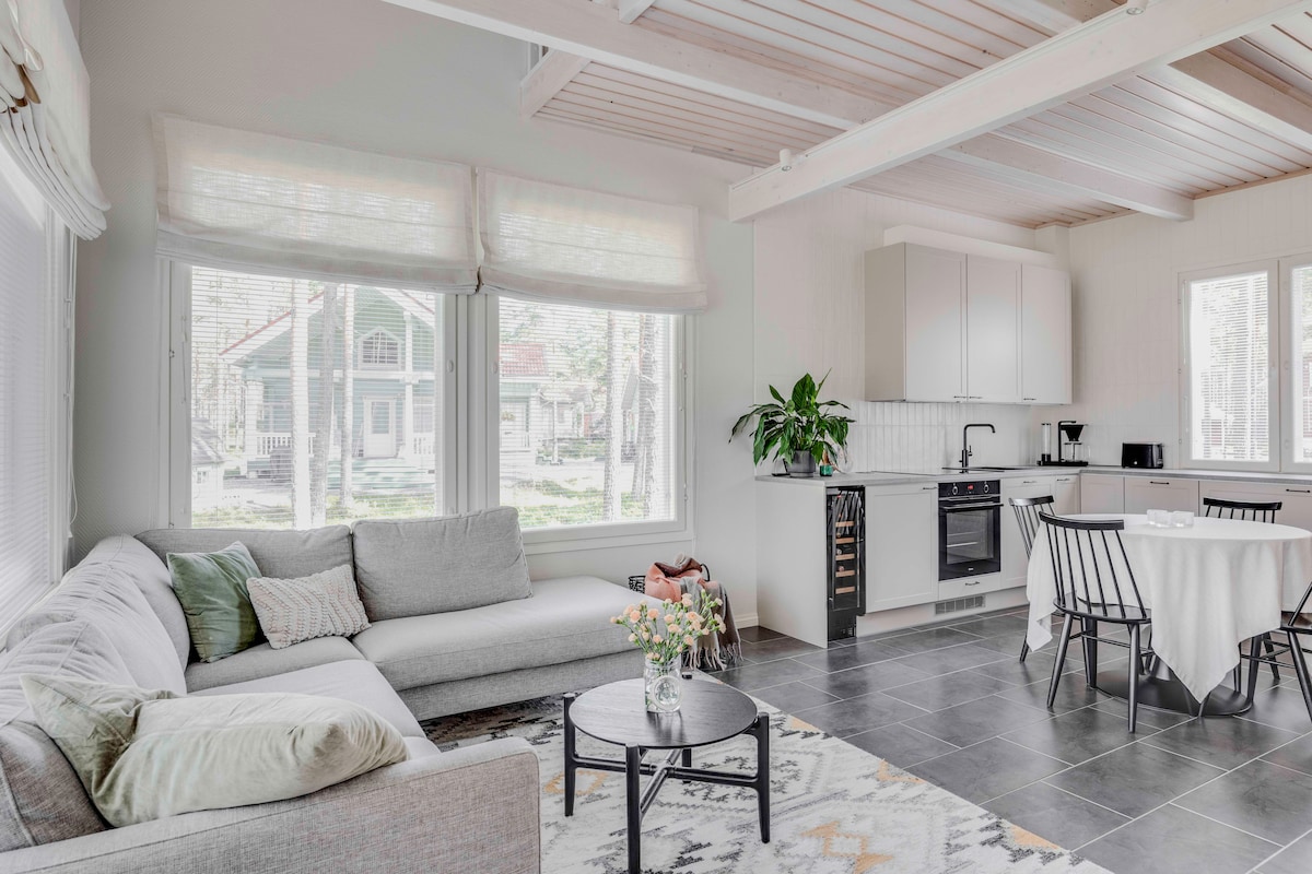 The living area features a spacious gray sectional sofa adorned with soft cushions. A round coffee table is placed in the center, surrounded by a light area rug. Large windows allow natural light to fill the room, with a view of the outdoors and a dining table set nearby.