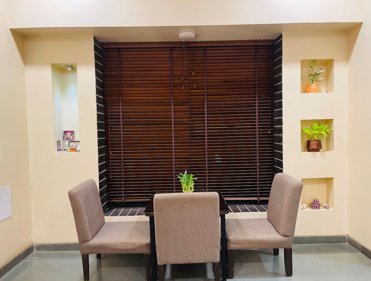 A small dining area is presented, featuring a dark wooden table surrounded by two light-colored chairs. Natural light is filtered through wooden blinds, creating a warm ambiance. Decorative shelves with plants and personal items are seen on the wall, adding a touch of greenery.