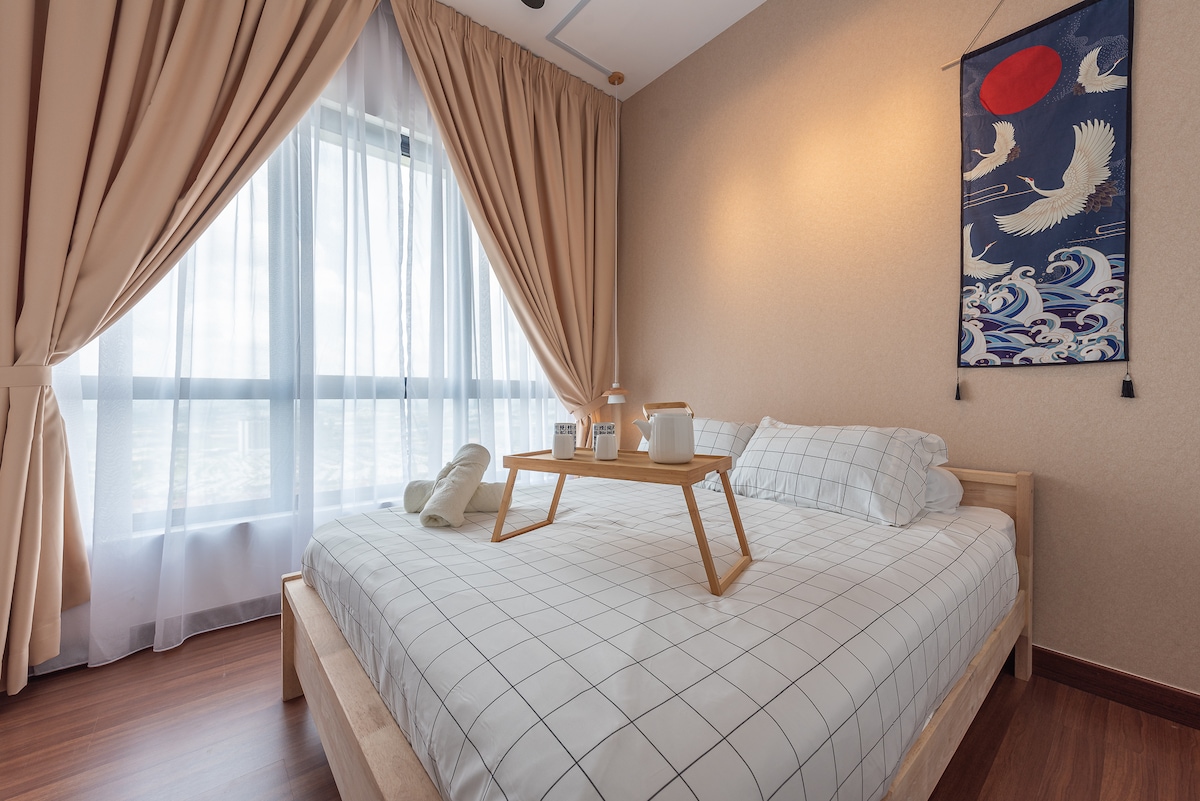 A spacious bedroom features a large bed dressed in a grid-patterned sheet. Natural light filters through sheer curtains, enhancing the neutral tones of the room. A wooden tray with two cups and bath towels is positioned on the bed, adding a functional touch to the inviting space.
