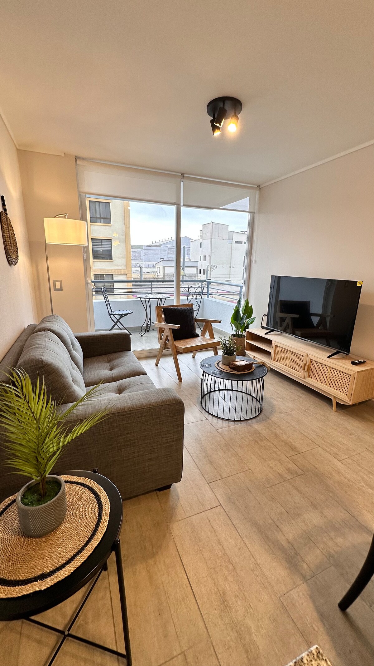 A cozy living area is presented with a gray sofa and wooden armchairs surrounding a circular coffee table. A television sits on a light wood media console beside a small indoor plant. Large windows provide natural light and views of the street.