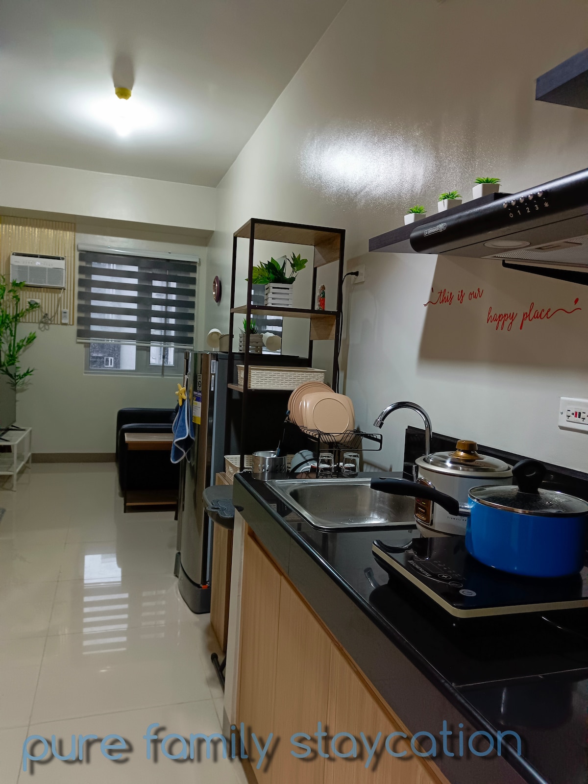 A compact kitchen area is equipped with essential appliances, including an induction stove and refrigerator. Counter space is highlighted with cookware and an electric kettle. A small dining area is visible through an open layout, complemented by natural light from nearby windows.