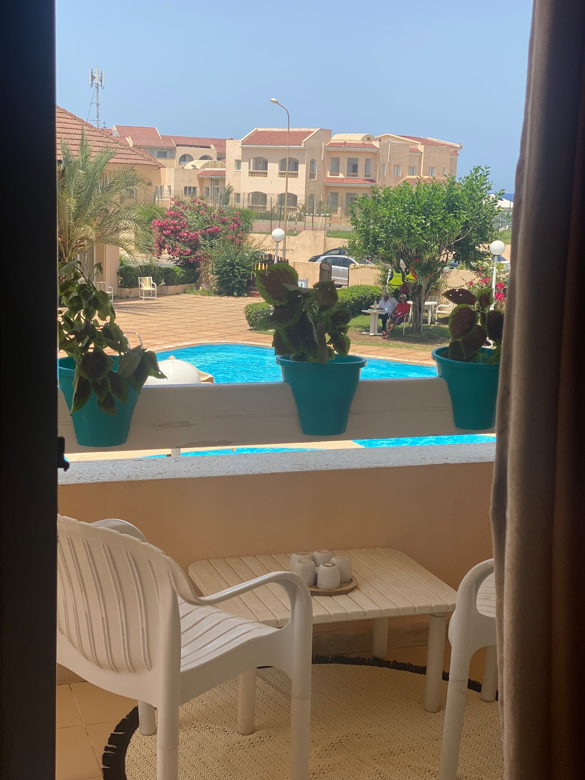 A balcony view reveals a serene pool area framed by vibrant greenery and colorful flowers. Potted plants adorn the railing, while two white chairs and a small table are positioned on the tiled floor, creating a peaceful spot to relax.