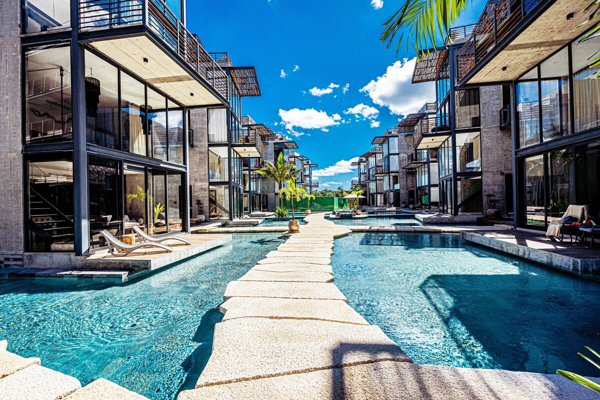 A serene landscape showcases modern apartments arranged around a central pool area. Stone bridges cross over reflective waters, flanked by palm trees. Bright blue skies and scattered clouds create a vibrant backdrop, inviting relaxation in a tranquil outdoor space.