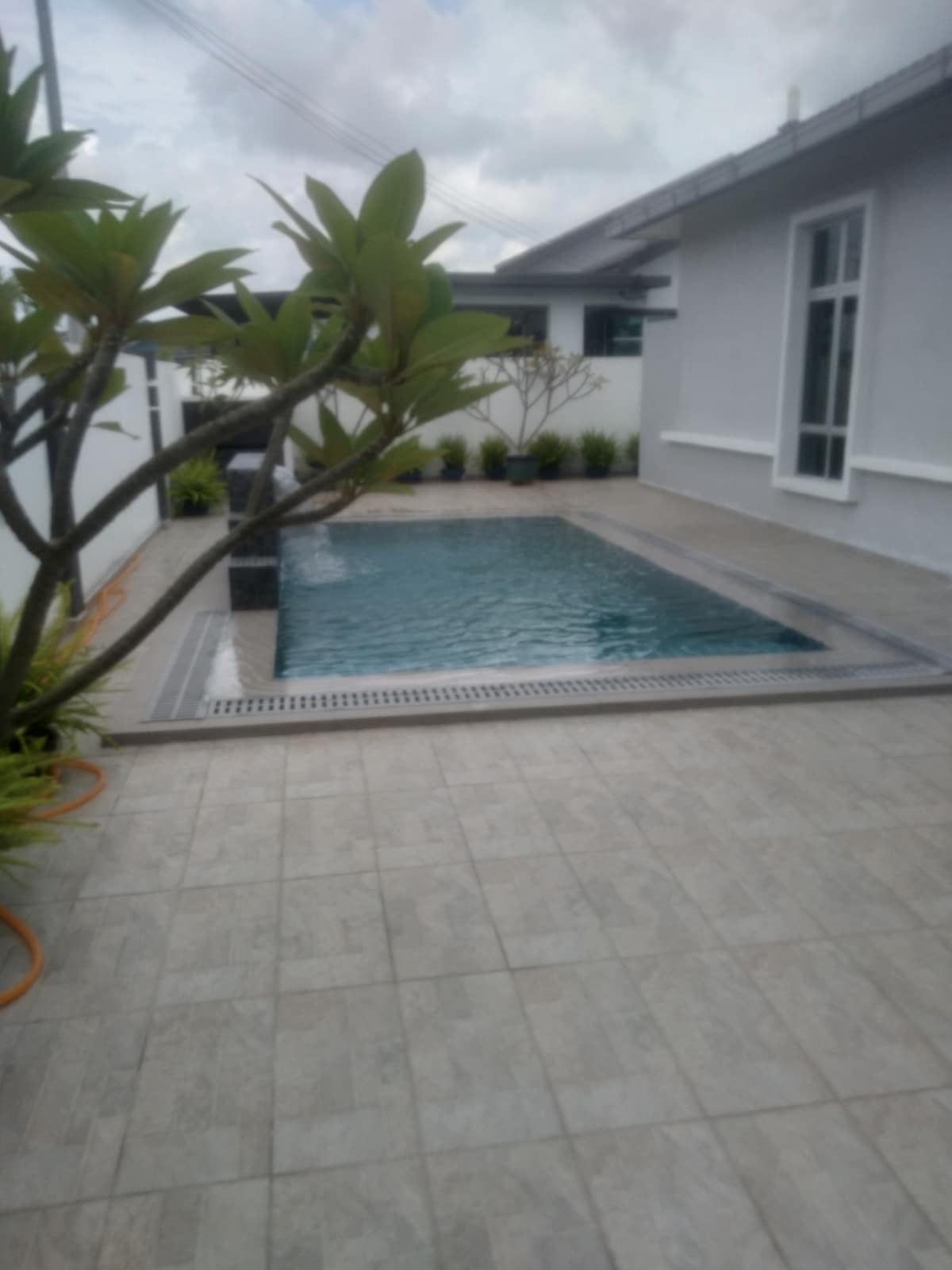A private swimming pool is surrounded by neatly arranged tiles, with greenery positioned along the edges. The area features a calm water surface reflecting the sky, while plants add an element of nature against the white walls of the property.