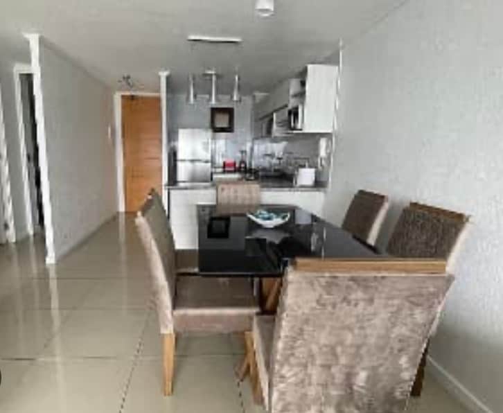 A dining area features a sleek black table surrounded by four beige upholstered chairs. In the background, a modern kitchen is visible, equipped with stainless steel appliances and a clean, minimalist design. Neutral-toned walls and tiled flooring create a spacious ambiance.