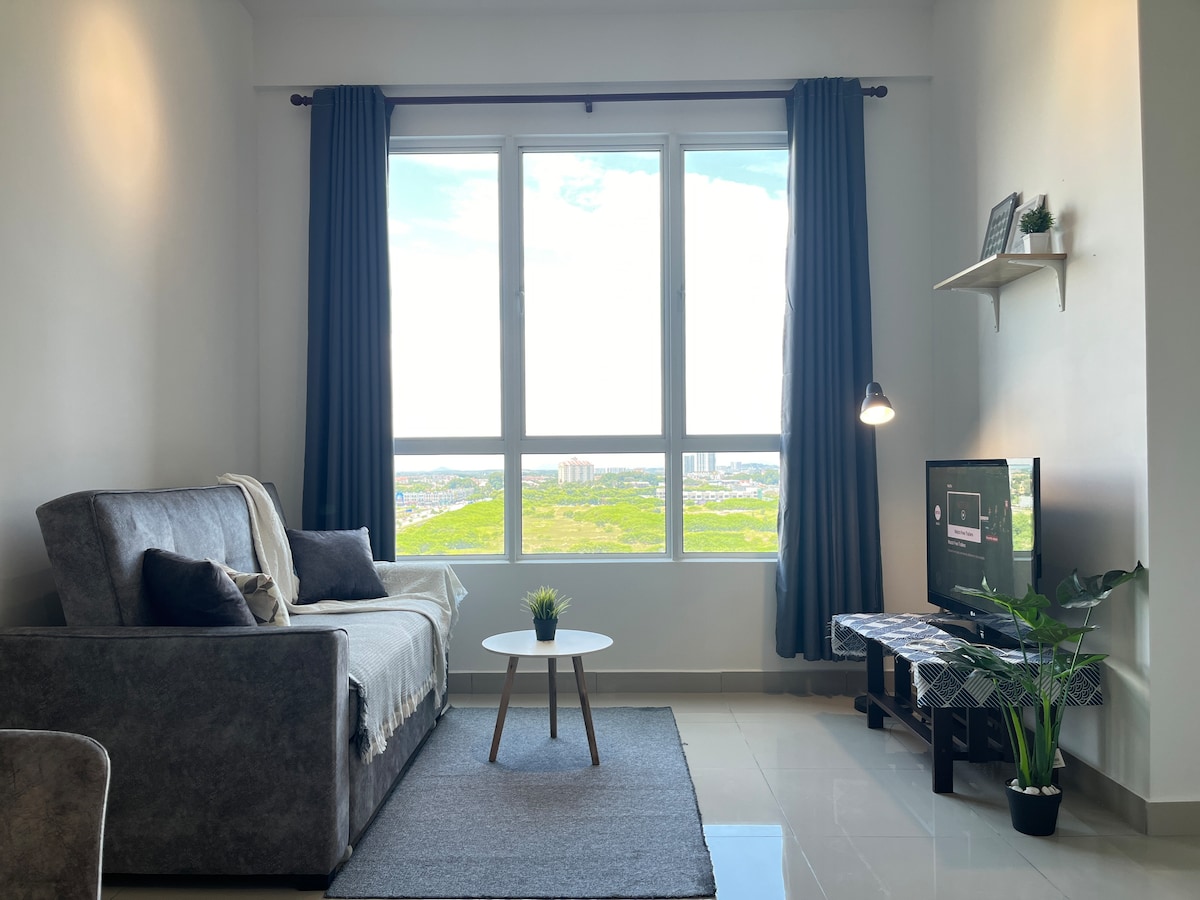 A comfortable living area is highlighted by a queen size sofa bed adorned with a light throw. A small round table sits on a grey rug, and a television is positioned next to a decorative plant. Large windows offer a view of the surrounding city landscape.