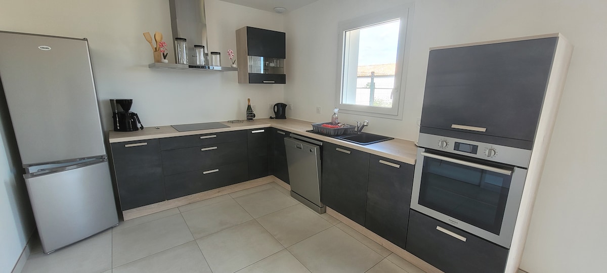 A modern kitchen is presented with sleek cabinetry in dark tones. Stainless steel appliances, including a refrigerator and an oven, are featured. A spacious countertop and a sink are integrated into the layout, with natural light flooding through the window.