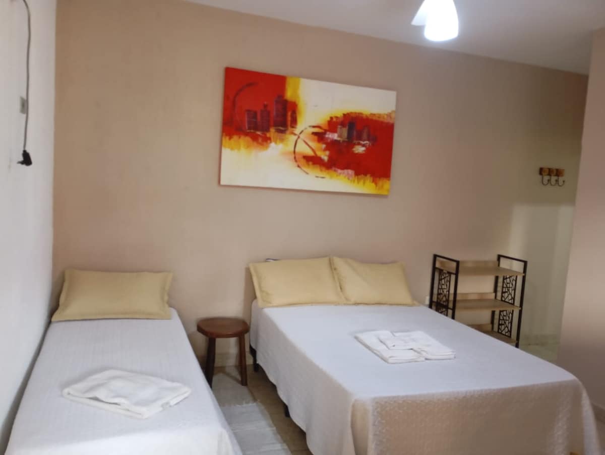 The room features two spacious beds, each topped with light-colored linens and decorative cushions. A colorful wall art piece adds visual interest above the beds. A small wooden table and a shelving unit are positioned to provide practical space for personal items.