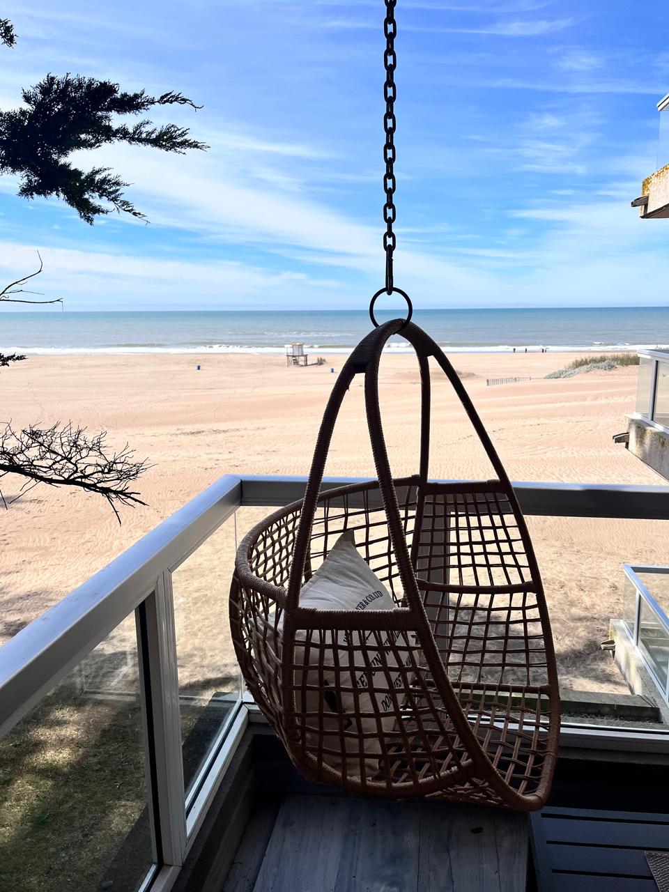 A hanging wicker chair is featured on the balcony, providing a view of the sandy beach and ocean. The clear sky enhances the serene landscape, while distant figures can be seen enjoying the beach. Natural light fills the space, creating a relaxed atmosphere.