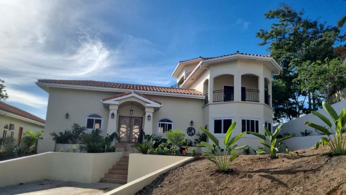 A charming two-story home is featured, with a welcoming entrance framed by lush greenery. The exterior showcases a light-colored façade with arched windows and a tiled roof, while a staircase leads up to the entrance, providing access to the inviting living spaces inside.
