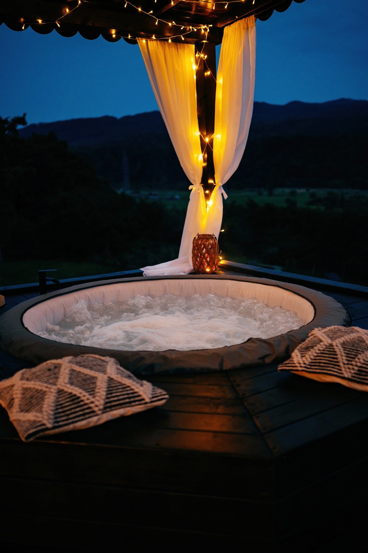 A hot tub is positioned under soft lighting, surrounded by a serene mountain backdrop. The warm water creates gentle bubbles, while decorative pillows are placed nearby for comfort. Sheer curtains drape elegantly, enhancing the tranquil atmosphere.