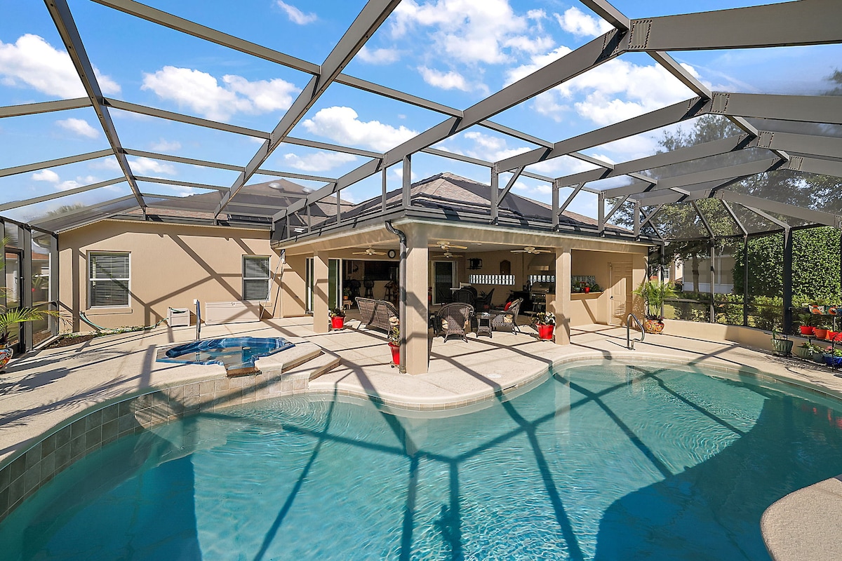 A spacious outdoor area is showcased, featuring a pool surrounded by a screened enclosure. Comfortable seating areas are visible, offering relaxation options. A hot tub is positioned next to the pool, with lush greenery and sky views enhancing the setting.