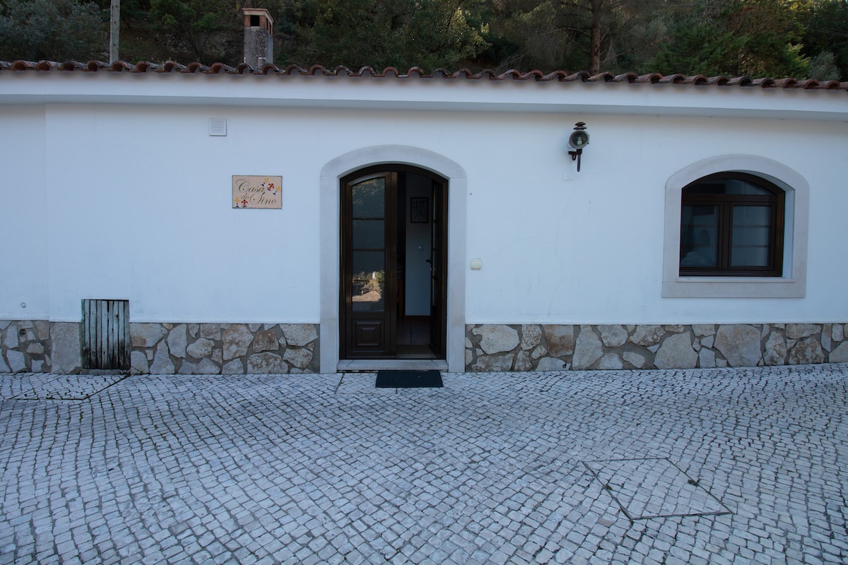 An entrance to a building is framed by a smooth stone facade and a rustic tiled roof. The doorway features a dark wooden door and a small decorative sign. Two arched windows with white frames flank the entrance, providing natural light to the interior.
