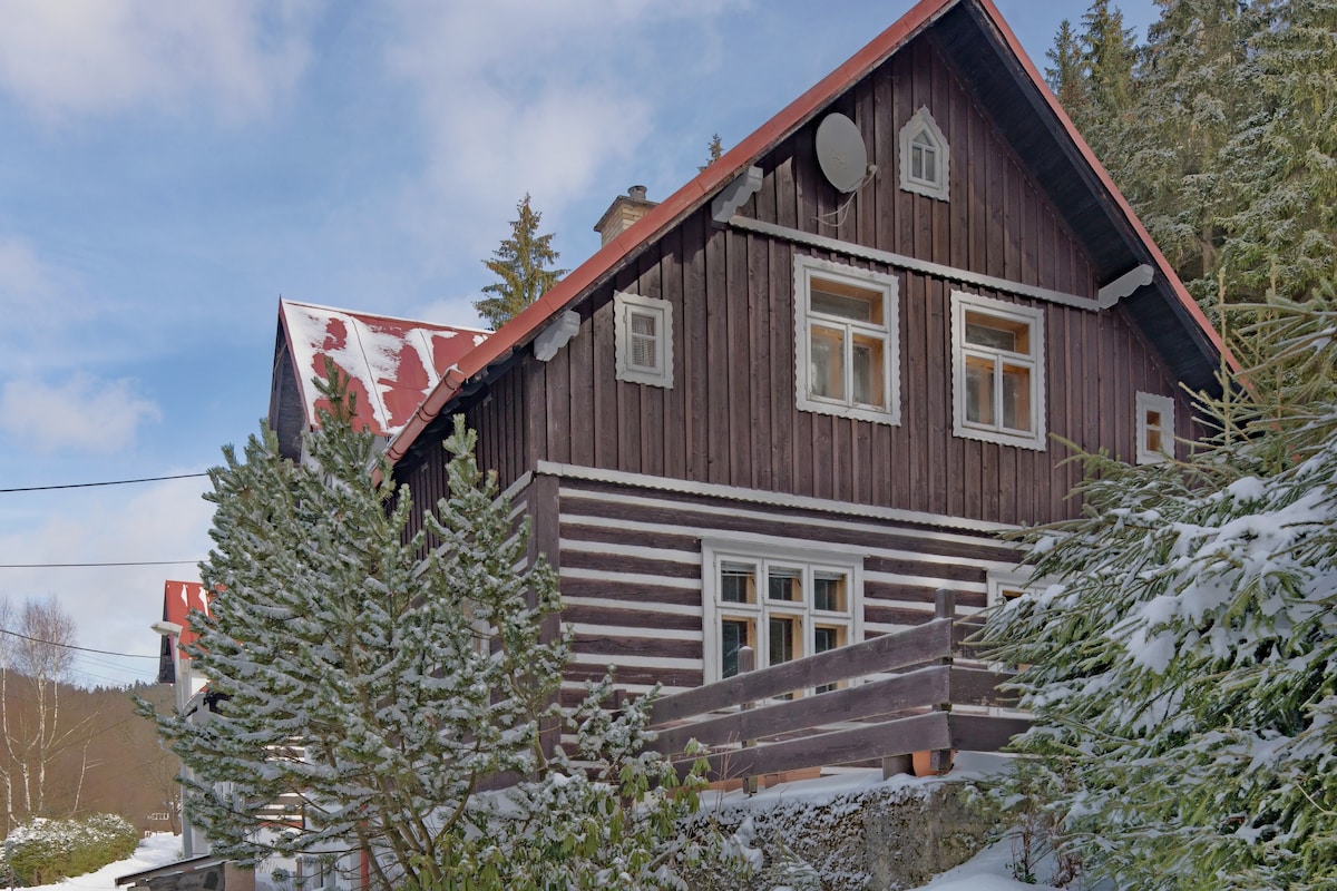 A charming wooden cabin is surrounded by snow-covered trees, with a distinctive red and white roof. Large windows reflect natural light, while a small balcony is visible above a wooden railing. The peaceful setting is nestled among the mountains, providing a sense of tranquility.