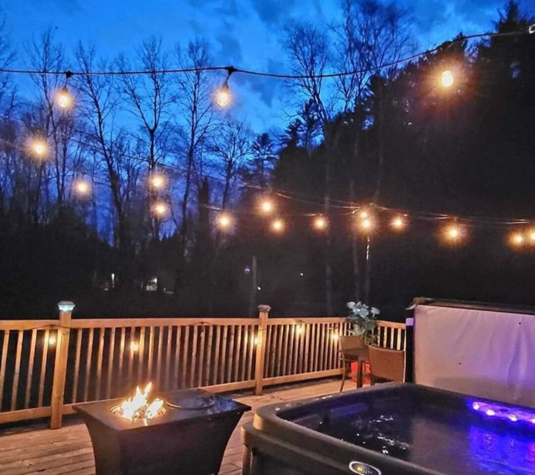 A deck area is illuminated by string lights, creating a warm glow against the twilight sky. A hot tub is visible alongside a fire pit, where flames flicker softly. Trees frame the scene, adding to the secluded setting.