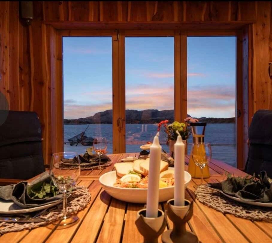 A dining table is set for a meal, featuring plates of food, glassware, and candles. Large windows frame a scenic view of the water and surrounding landscape, with soft natural light enhancing the warm wooden interior.