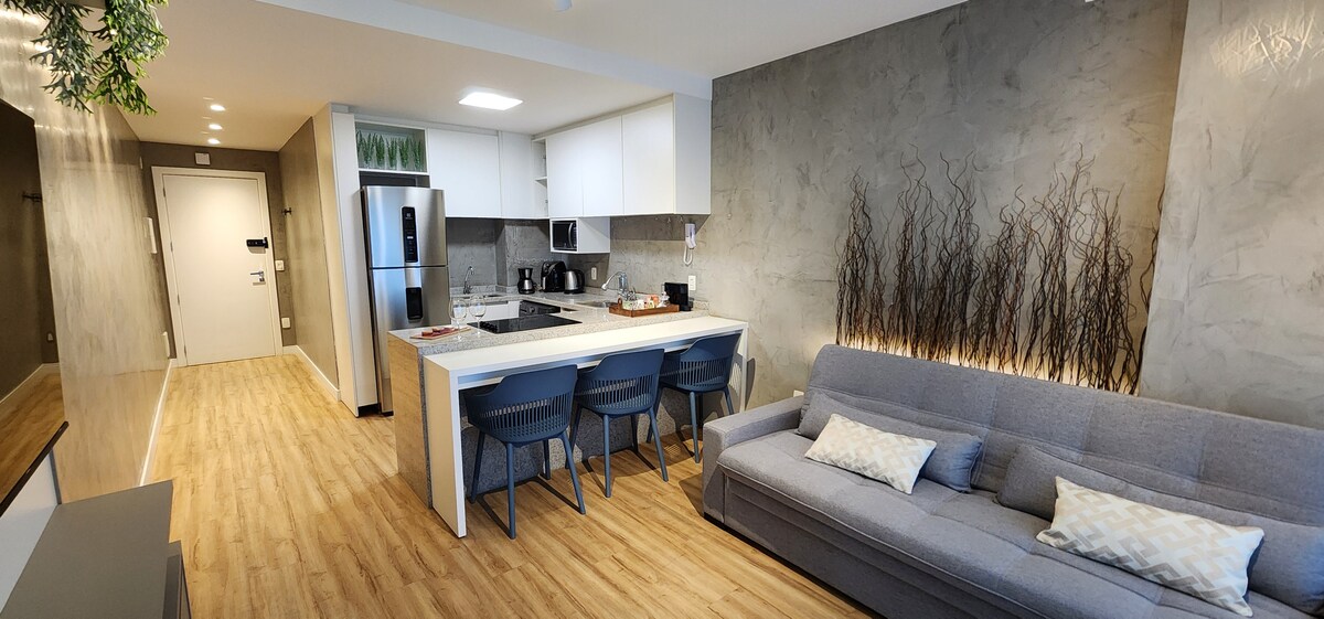A modern kitchen area is visible, equipped with stainless steel appliances and a white countertop. A grey sofa and three matching chairs around a dining table complete the inviting living space. The walls are adorned with natural elements, creating a serene environment.