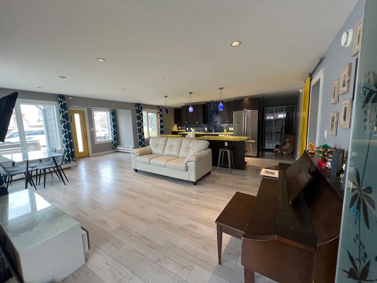 An open-plan living area is presented, featuring a light-colored sofa and a dark wood piano. The kitchen is visible in the background, equipped with modern appliances and a yellow-lit countertop. Windows allow natural light to fill the space, complemented by patterned curtains.