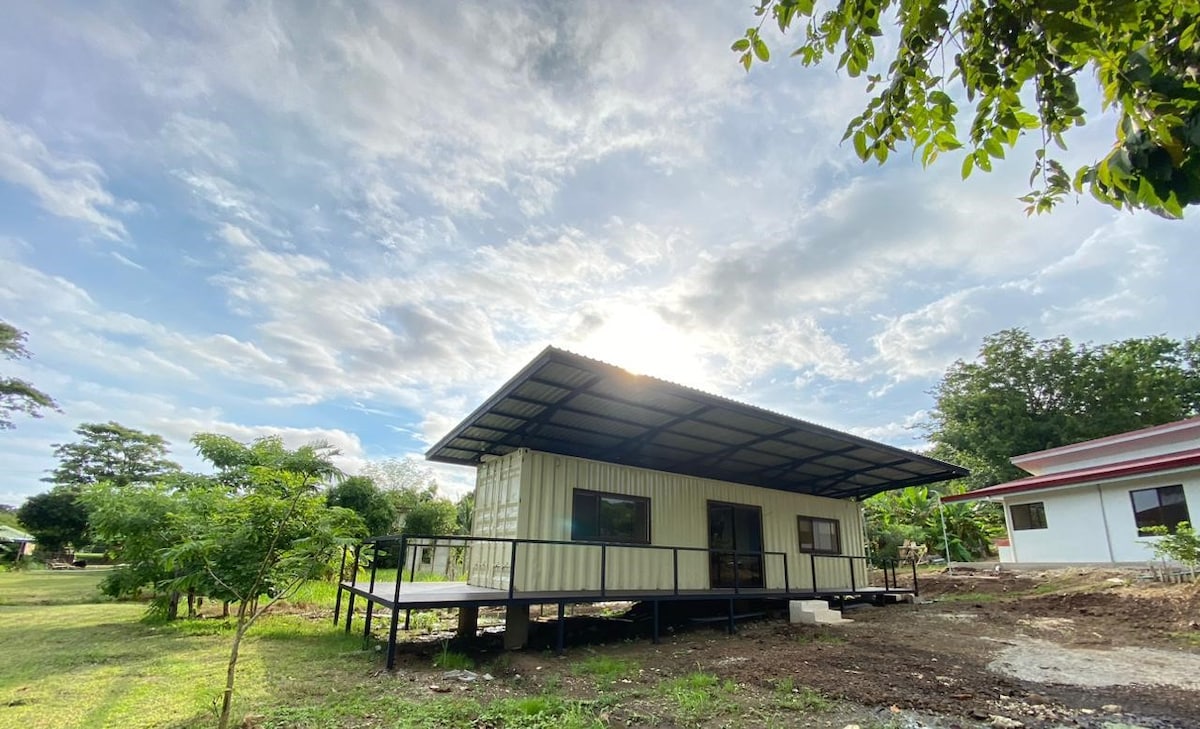 A modern structure with a metal roof is surrounded by lush greenery and open space. Large windows allow natural light to fill the interior, while a wooden deck provides an outdoor area for relaxation. The sky is partly cloudy, adding a serene atmosphere.