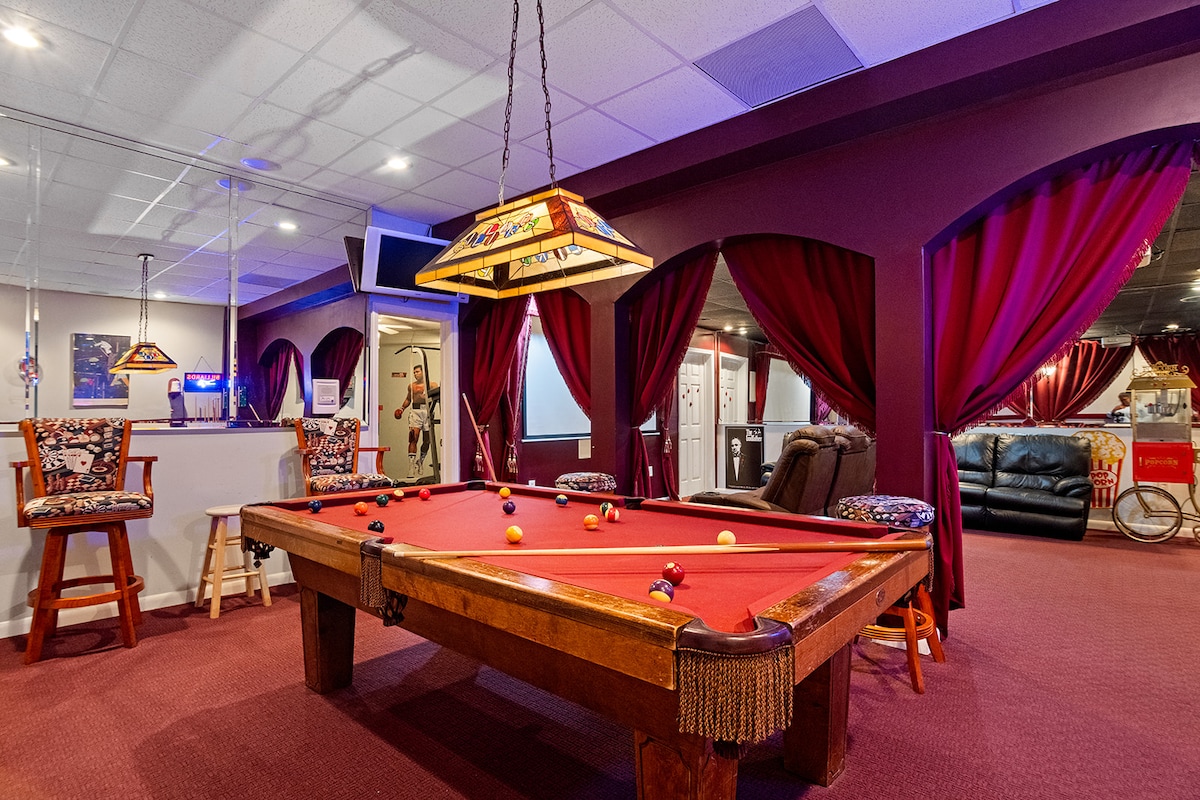 A finished basement entertainment area features a classic billiards table with a red felt surface. Surrounding the table, plush seating provides comfort. Rich burgundy curtains frame the space, creating a cozy atmosphere, complemented by soft overhead lighting.