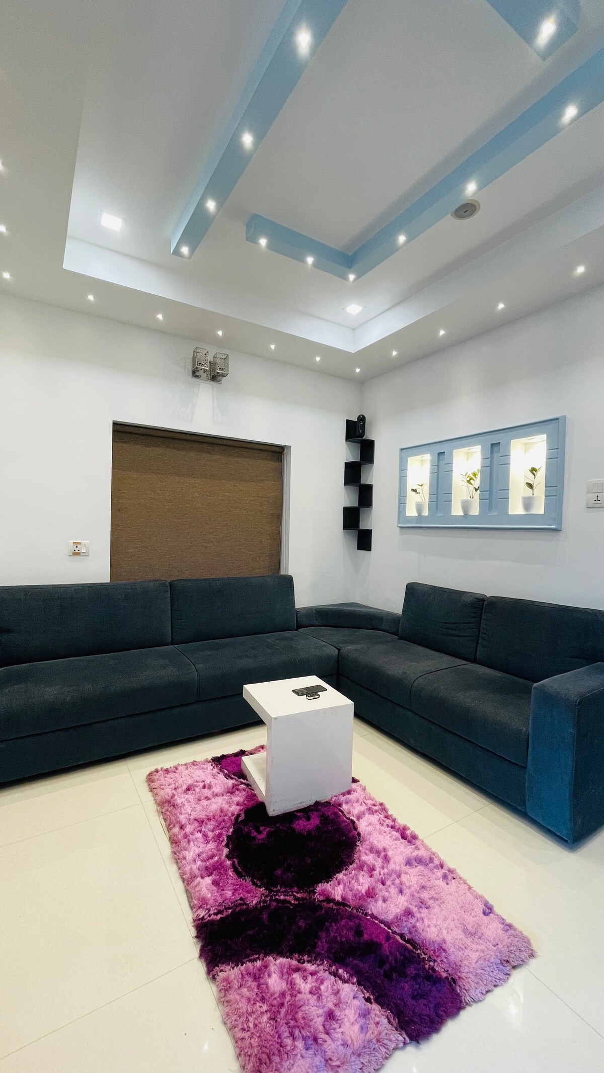 A modern living area features a dark blue sectional sofa arranged around a small white coffee table. A plush purple area rug adds a pop of color to the floor. Ambient lighting emanates from the ceiling, enhancing the inviting atmosphere.