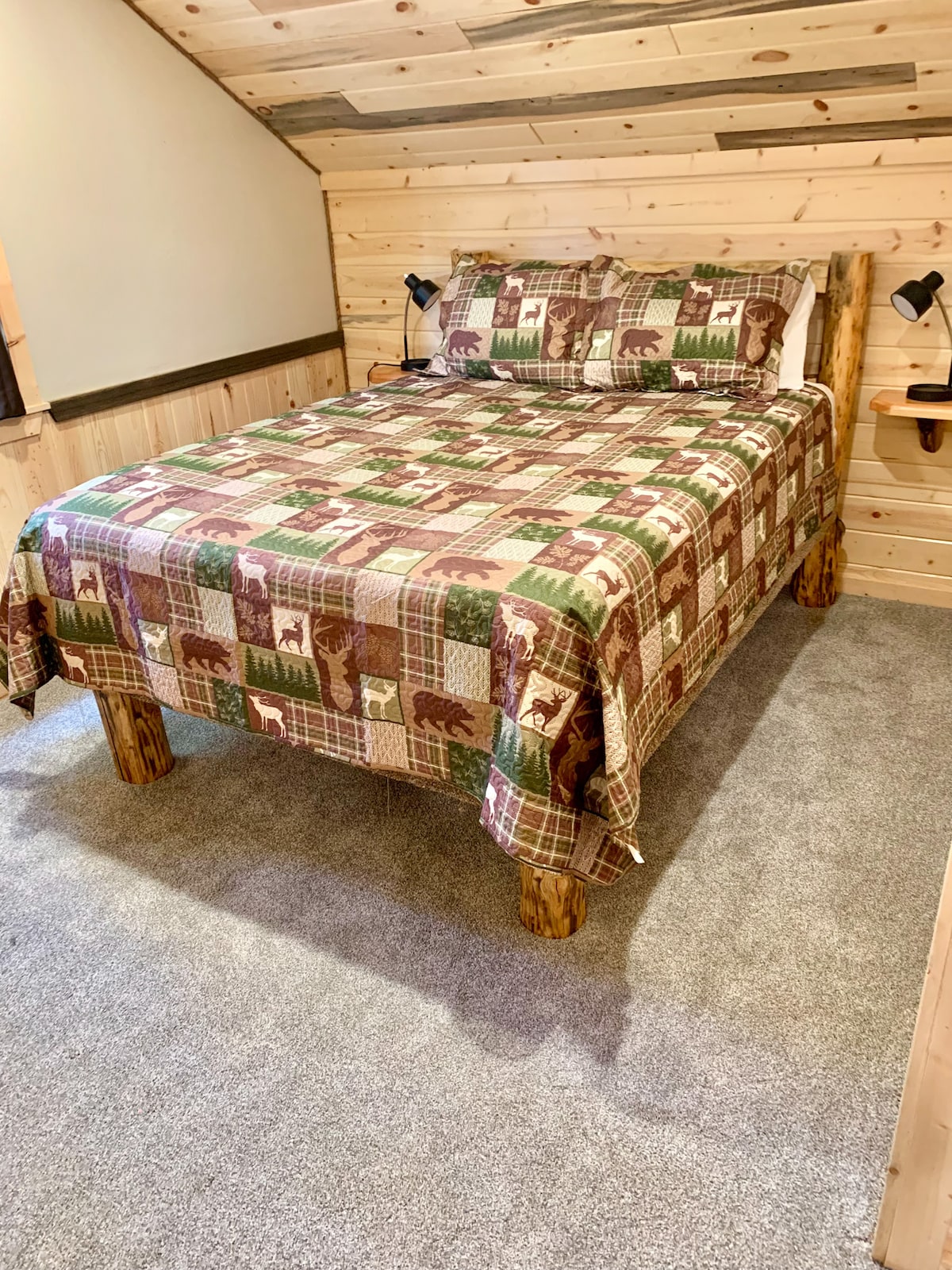A simple bedroom with a queen-size bed featuring cabin-style bedding and matching pillows. The room has carpeted flooring, wood-paneled walls, and small wall-mounted reading lamps for convenience. 