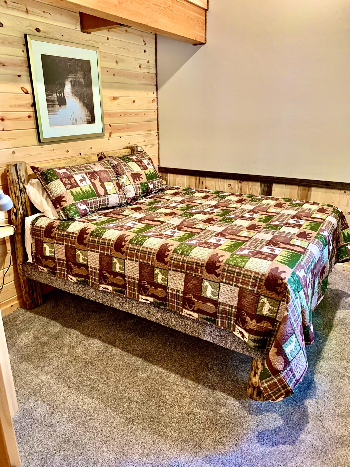 A simple and cozy bedroom featuring a queen-size bed with cabin-style bedding and matching pillows. The room has wood-paneled walls, carpeted flooring, and clean décor, offering a comfortable and functional sleeping space. 