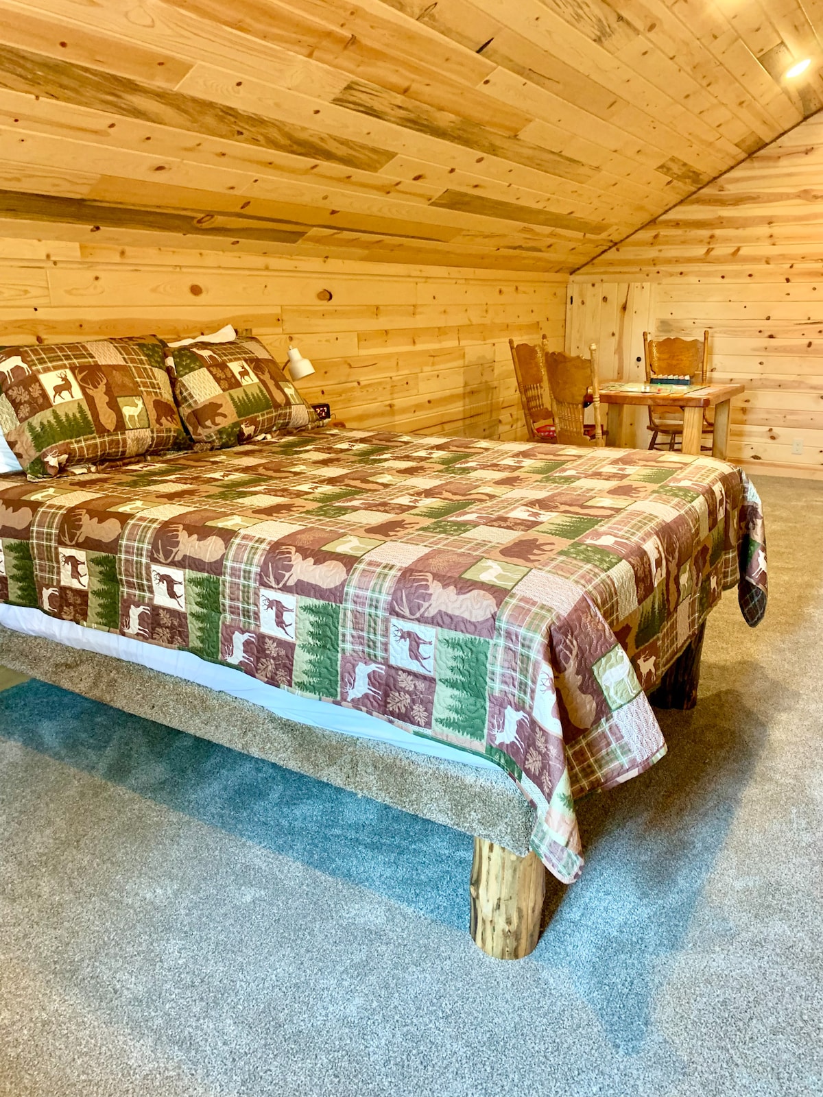 A cozy bedroom with a queen-size bed featuring cabin-style bedding and matching pillows. The room has carpeted flooring, wood-paneled walls, a bedside lamp, and a small table with chairs, creating a warm and simple cabin atmosphere. 