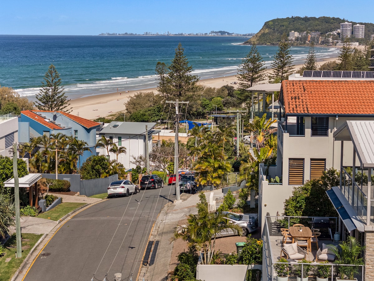 A panoramic view showcases the beachside landscape, with golden sands stretching along the coastline. Coastal trees frame the scene, while distant city structures can be seen against the ocean backdrop. The roadway below leads to the beach area, reflecting a serene seaside atmosphere.
