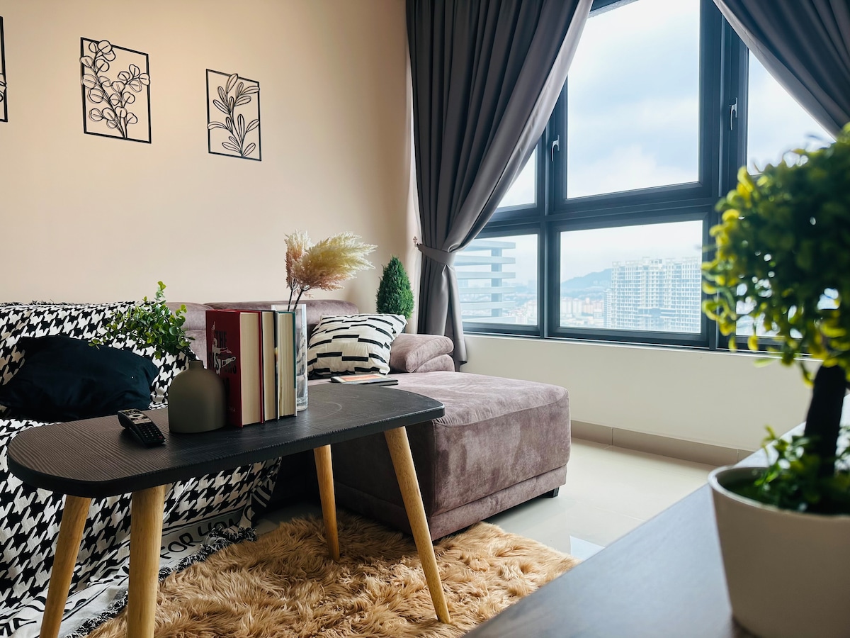 A modern living area is showcased, featuring a plush sofa with colorful cushions beside a low coffee table. Decorative plants and books are placed on the table, while large windows allow natural light to fill the space, revealing a view of the surroundings.
