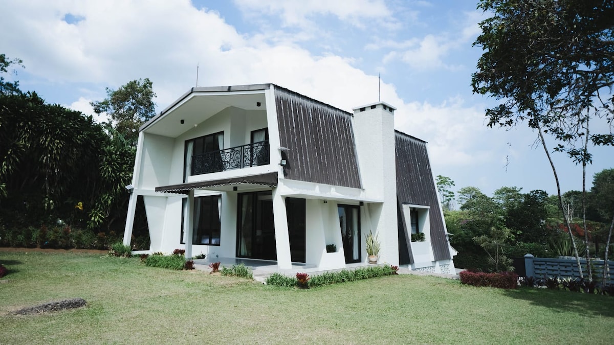 The villa is presented in a modern architectural style, featuring a sloped roof and large glass windows that allow natural light to fill the interior. Surrounding greenery and vibrant landscaping enhance the serene atmosphere, creating a welcoming outdoor space.