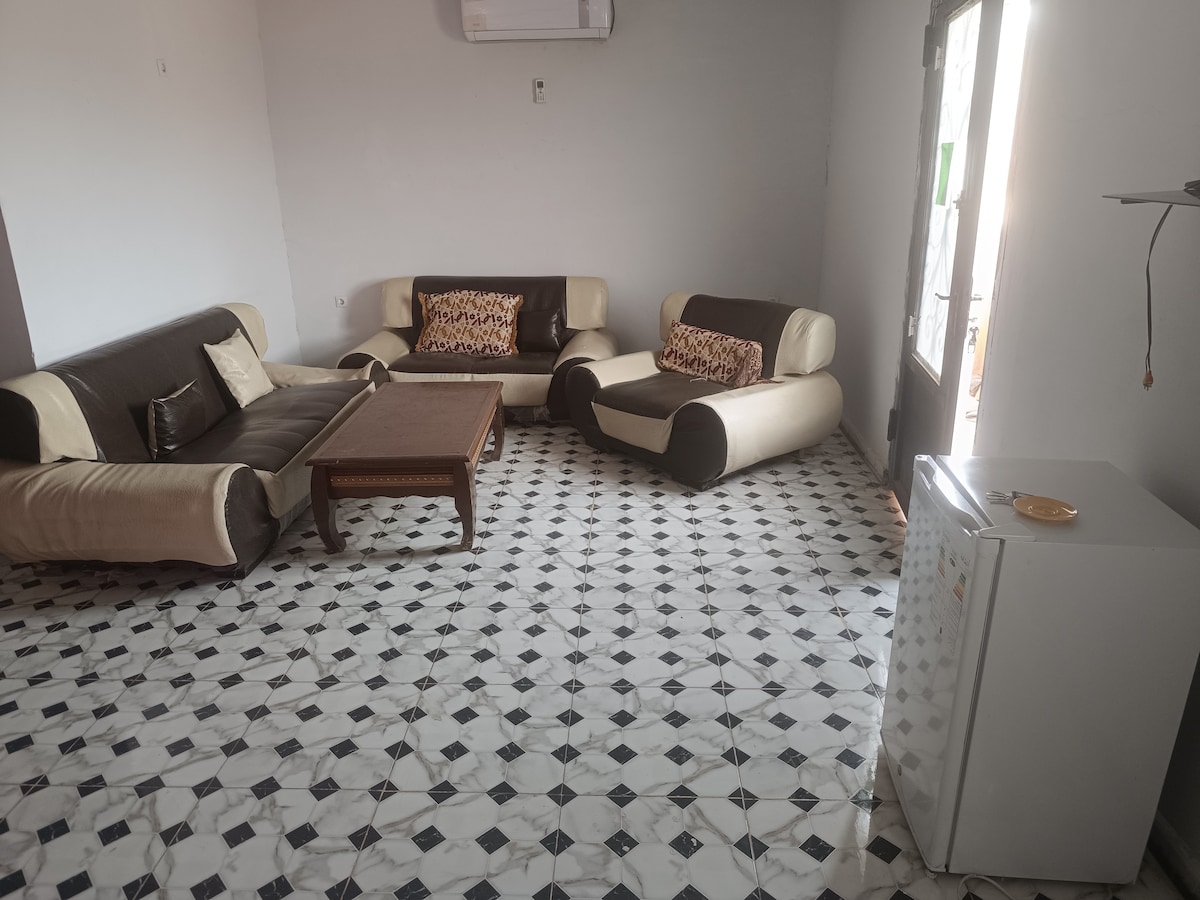 A cozy living area features a set of contemporary sofas with contrasting colors and a wooden coffee table at the center. The tiled floor showcases a black and white geometric pattern, while a small refrigerator is seen along one wall. Natural light enters through a nearby window.