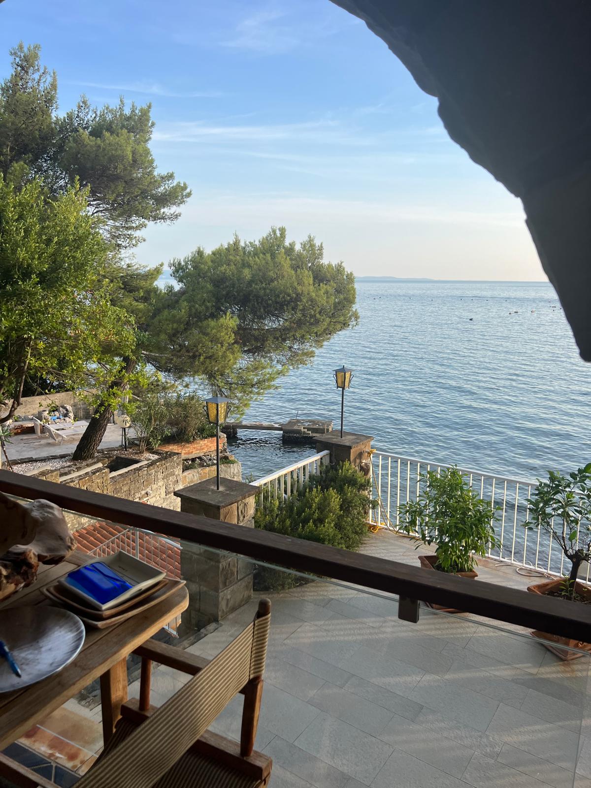 A serene view from a window showcases calm waters, gently lapping against the shore. Lush green trees are visible nearby, framing the scene. A cozy outdoor area with a stone pathway and potted plants enhances the tranquil atmosphere, inviting relaxation by the sea.